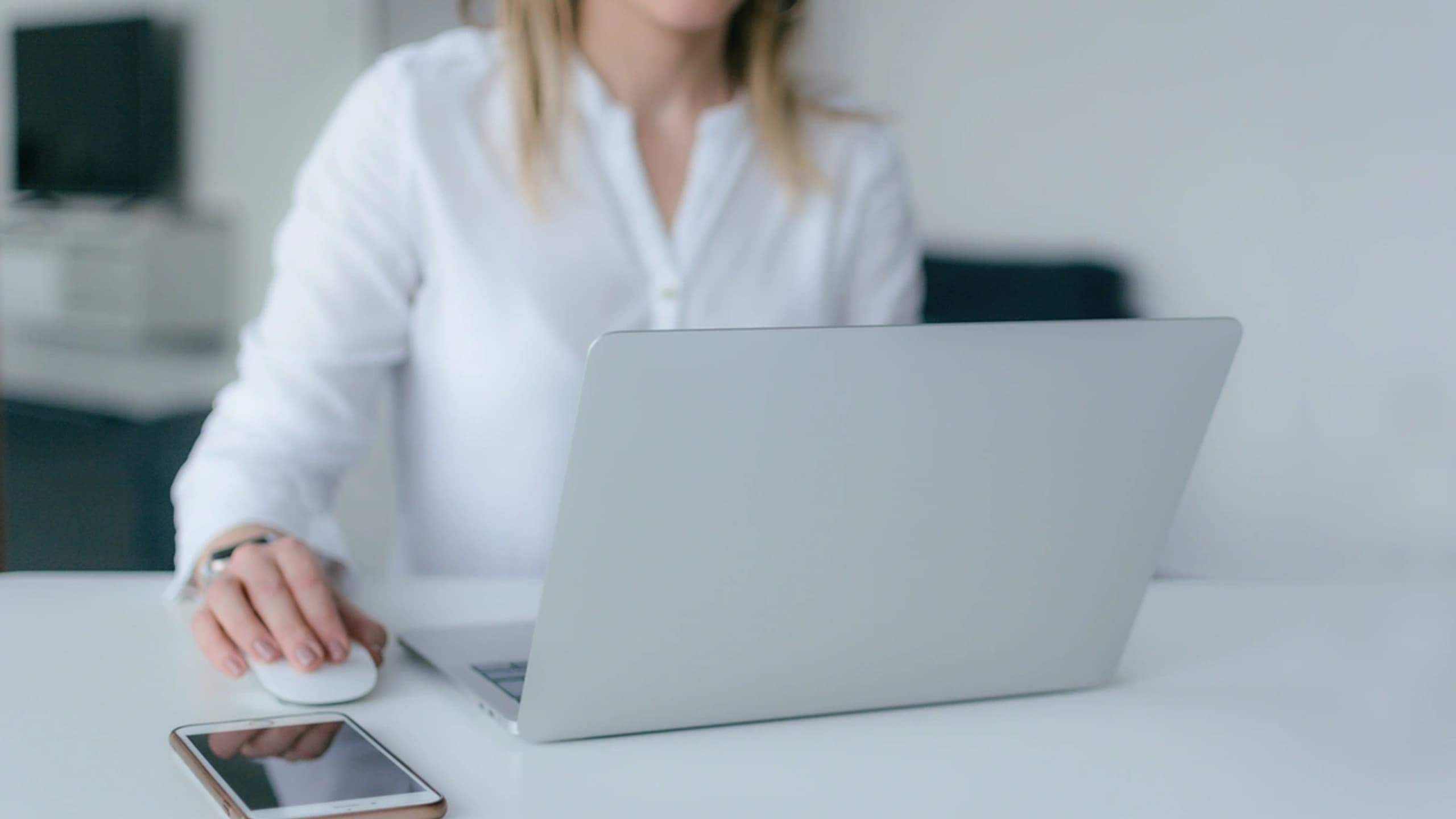 A woman is working at her computer.  Her phone is on the desk by her